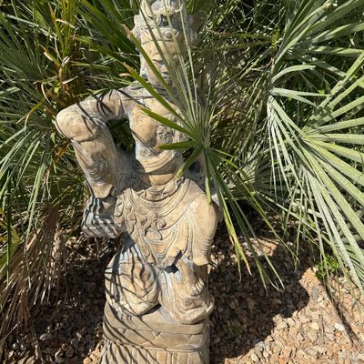 Sale Photo Thumbnail #39: Weathered stone garden statue depicting an Asian deity or Bodhisattva, positioned on a pedestal among foliage.