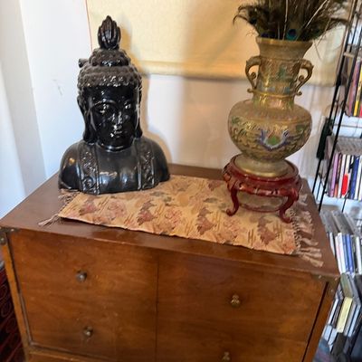 Sale Photo Thumbnail #13: Black ceramic Buddha head bust alongside a cloisonné brass vase with peacock feathers, displayed atop a campaign-style wooden chest.