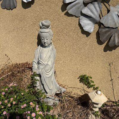 Sale Photo Thumbnail #40: Cast stone Guanyin garden statue standing in a garden bed alongside metal ginkgo leaf wall decor mounted on a stucco wall.