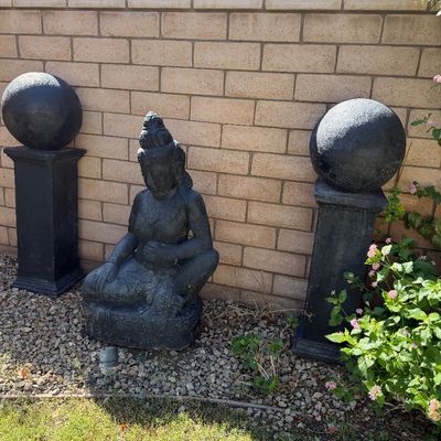 Sale Photo Thumbnail #35: Black stone garden Buddha statue flanked by a pair of matching stone pedestal sphere orb ornaments set against a brick wall.
