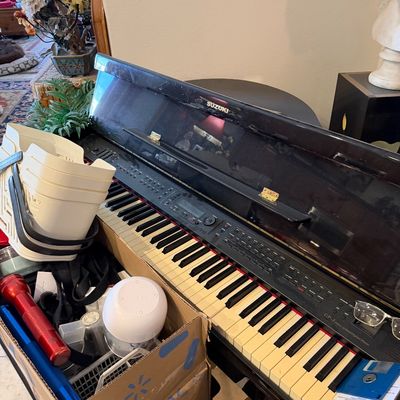 Sale Photo Thumbnail #19: Suzuki GP-3 Mini Grand digital piano with a black high-gloss finish, displaying light surface wear, accompanied by a box of miscellaneous household items.