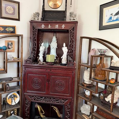 Sale Photo Thumbnail #3: Intricately carved Asian altar cabinet topped with a gong and flanked by open display shelving filled with various figurines and decor.