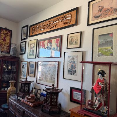 Sale Photo Thumbnail #24: Collection of Asian art and decor including framed Japanese ukiyo-e woodblock prints, carved wooden wall panels, miniature pagodas, and a geisha doll in a glass display case.