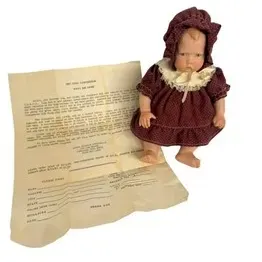 Vintage small baby doll dressed in a burgundy bonnet and lace-trimmed dress, accompanied by original paper documentation.