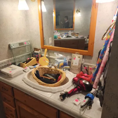 Bathroom vanity cluttered with various household items, personal care electronics, a wicker basket filled with accessories, and rolls of...