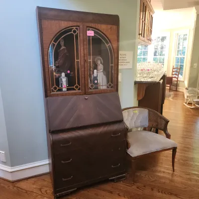 Vintage dark wood secretary desk with glass-front display cabinet and lower drawers, shown alongside a low tufted barrel chair.