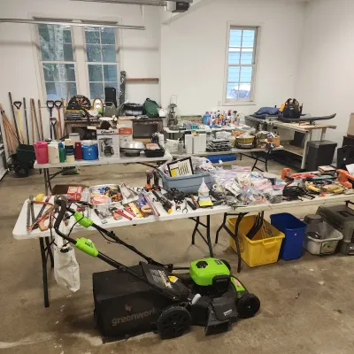 Sale Photo Thumbnail #127: A Greenworks electric lawn mower in the foreground surrounded by folding tables filled with various tools, yard equipment, and household goods inside a garage.