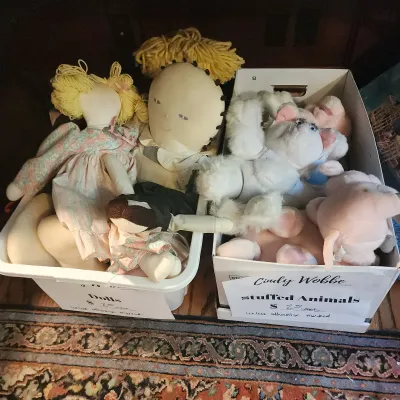 Two storage bins containing assorted vintage rag dolls with yarn hair and plush stuffed animals.