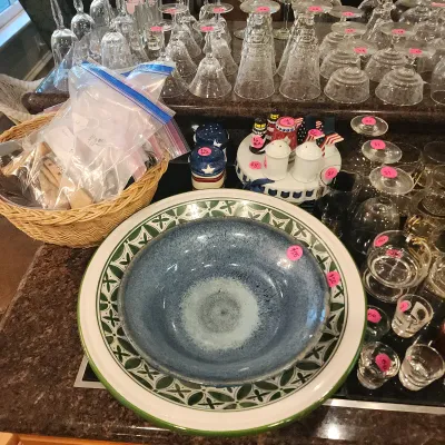 Large blue glaze ceramic bowl nested in a green geometric pattern serving bowl, surrounded by glassware, a woven basket, and novelties.