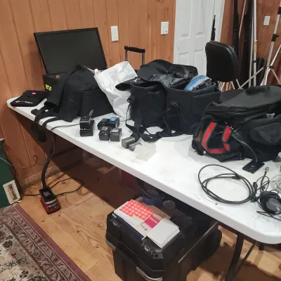 Sale Photo Thumbnail #148: Assortment of photography and video gear including soft camera bags, batteries, chargers, cables, a monitor, and tripods arranged on a folding table.