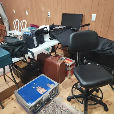 Sale Photo Thumbnail #158: Collection of equipment cases and bags, featuring a vintage brown leather suitcase, a blue flight case, camera bags, and a black office chair in front of a folding table.