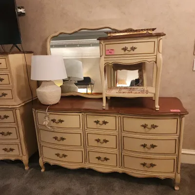 French Provincial style cream and wood-tone dresser set with matching wall mirror, nightstand, and textured ceramic table lamp.