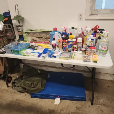 Sale Photo Thumbnail #131: White folding utility table displaying assorted household cleaning supplies, lawn edging, and hardware items alongside a green canvas bag and blue foam mat.