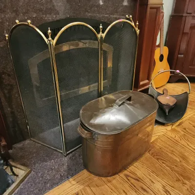 Brass-trimmed three-panel arched mesh fireplace screen paired with a vintage oval copper boiler tub and a scuttle holding a wooden...