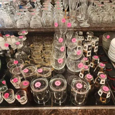Sale Photo Thumbnail #126: Large assortment of vintage glassware, including wine stems, champagne flutes, shot glasses, and gold-trimmed barware, marked with individual price tags.