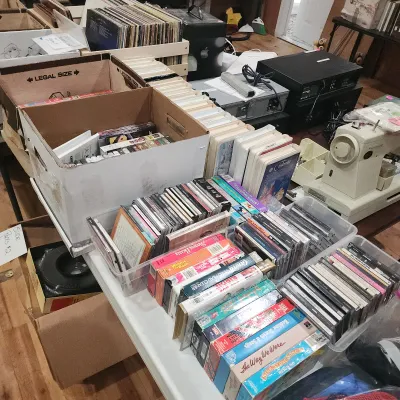 Collection of vintage media including vinyl records, VHS tapes, and CDs alongside home audio equipment and a Kenmore sewing machine.