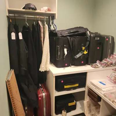 Closet area filled with tagged clothing on hangers, various soft and hard-sided luggage, and assorted craft supplies.