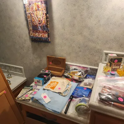 A collection of household items, perfumes, and electronics arranged on a bathroom counter, alongside a small decorative tapestry wall...