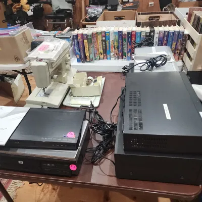 Collection of home electronics and media including DVD players, a vintage portable sewing machine, and a row of classic Disney VHS tapes...