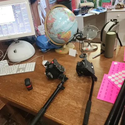 Cluttered wooden desk featuring a vintage Apple iMac G4 computer, a desktop world globe, camera tripod heads, and various office...