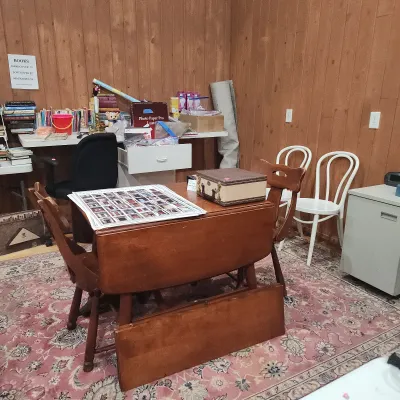 Vintage drop-leaf wooden dining table with matching chairs, set atop a floral area rug in a room filled with books and household supplies.