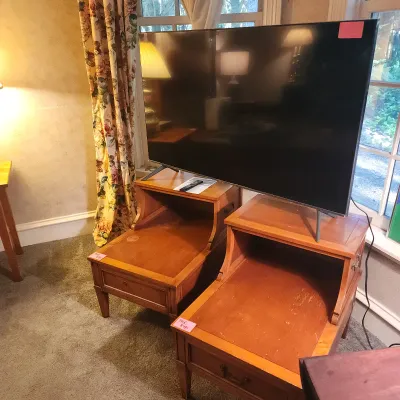 Pair of traditional mid-century style wooden step-up end tables supporting a large flat-screen television with a remote control.