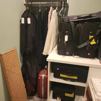 Collection of luggage pieces, vintage coats, and a white knit sweater in a closet setup alongside a folding sewing pattern board.