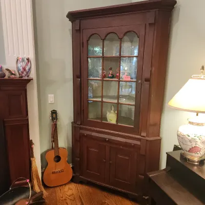 Antique corner hutch featuring arched nine-pane glass door top cabinet above a two-door lower cabinet. Acoustic guitar leaning nearby.