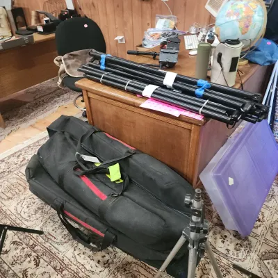 Collection of photography gear including lightweight light stands, a tripod, soft carrying bags, and office furniture in the background.