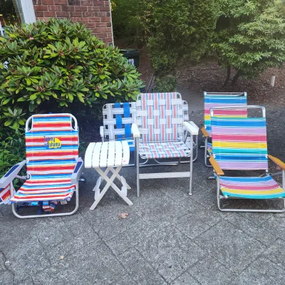 Collection of outdoor beach and lawn furniture including colorful striped folding chairs, webbing chairs, and a small white slatted...