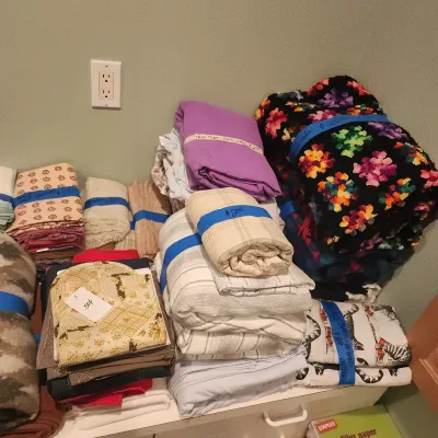 Assortment of vintage folded linens, blankets, and a handmade colorful crochet Afghan quilt, priced individually.