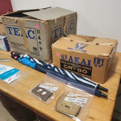Sale Photo Thumbnail #141: Vintage audio equipment boxes including a TEAC stereo tape deck box and an Akai 8-track cartridge stereo recorder box, alongside an umbrella and bagged outlet covers on a wooden workbench.