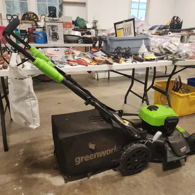 Greenworks 40V cordless lawn mower with attached rear grass catcher bag, displayed in a garage setting with various household tools in...