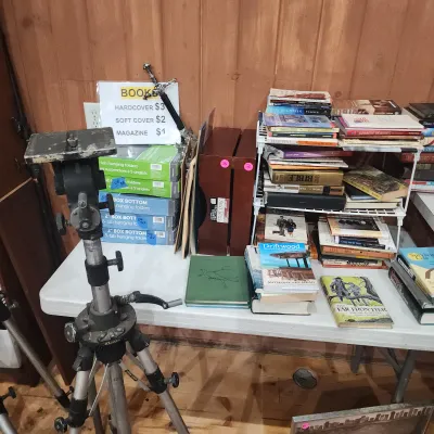 Sale Photo Thumbnail #154: A vintage heavy-duty metal tripod set up next to a table displaying stacks of books, office supplies, and decorative boxes.