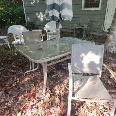 Outdoor patio set featuring a metal-framed glass top table with umbrella, beige mesh armchairs, and white wicker chairs in a yard.