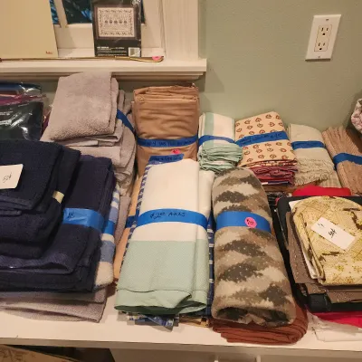 Assorted household linens including bath towels, table runners, tab-top curtains, and quilted potholders organized with price tags.