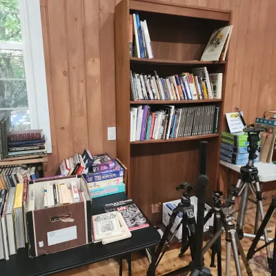 Sale Photo Thumbnail #147: Tall wood bookcase filled with assorted books, accompanied by additional books, board games, and photography tripods on surrounding tables.