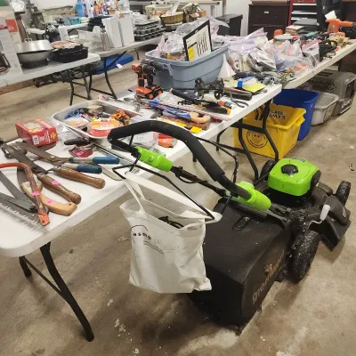 Greenworks electric push lawn mower surrounded by various hand tools, power tools, and hardware displayed on folding tables.