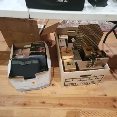 Two cardboard storage boxes filled with photography equipment, including vintage Nikon camera lenses in original gold boxes, a Polaroid...