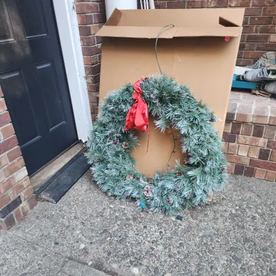 Frosted artificial Christmas wreath adorned wit
h pinecones and a red velvet bow, resting against its original storage box.