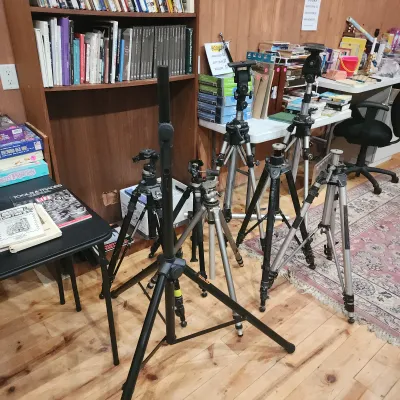 Sale Photo Thumbnail #153: Collection of vintage camera and lighting tripods in various sizes and finishes, arranged on a wood floor in front of bookshelves.
