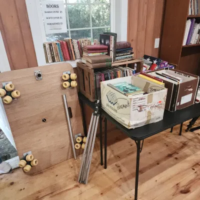 Sale Photo Thumbnail #146: Collection of vintage books displayed on tables and in crates alongside a wooden dolly board, metal camera tripod, and storage boxes.