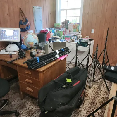 Home office filled with photography equipment, including various tripod stands, lighting gear, black equipment bags, an Apple iMac...