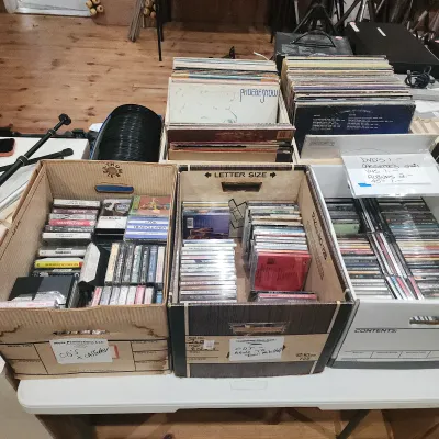 Large collection of media including vinyl records, cassette tapes, and CDs organized in cardboard boxes.