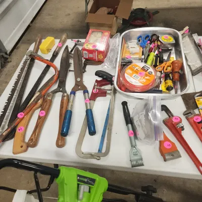 Sale Photo Thumbnail #136: Assortment of hand tools on a table, including hedge shears, hand saws, pipe wrenches, and a aluminum tray filled with scissors and tape measures.