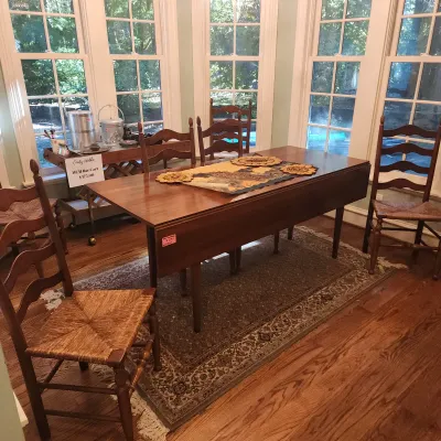 Sale Photo Thumbnail #115: Traditional wooden drop-leaf dining table paired with a set of ladderback chairs featuring rush woven seats, displayed on a textured area rug in a sunroom.