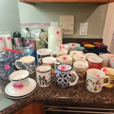 Collection of assorted ceramic mugs, glassware, and kitchenware displayed on a countertop, individually priced with pink sticker tags.