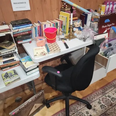 Sale Photo Thumbnail #155: Black ergonomic rolling office chair positioned in front of a white desk filled with books, small knick-knacks, and household supplies.