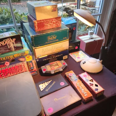 Collection of vintage board games, handheld electronic games, and domino sets arranged on a table under a desk lamp.