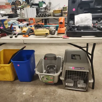 Sale Photo Thumbnail #130: Assorted power tools including drills, sanders, and a Craftsman tool set arranged on a table above plastic bins and a Petmate Kennel Cab crate.